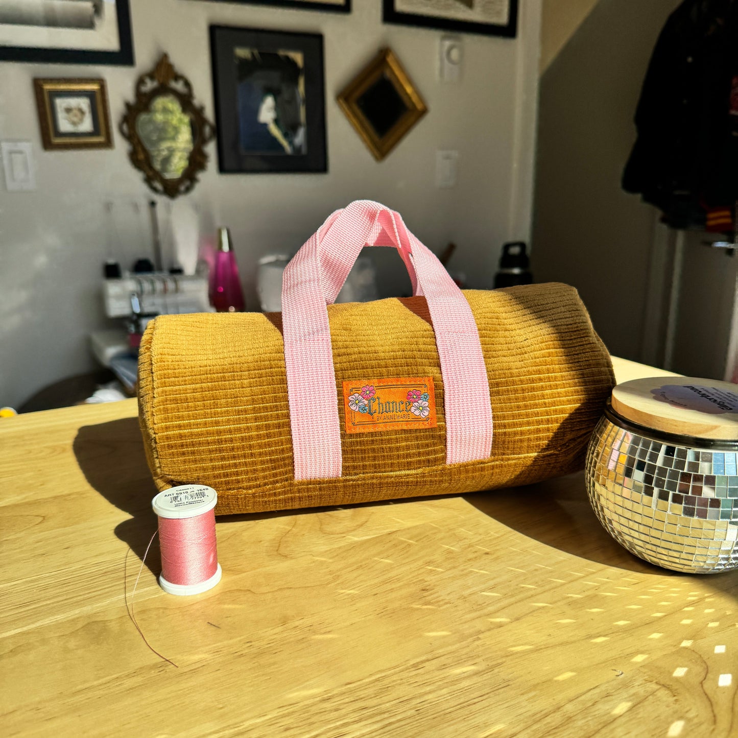 A mustard yellow corduroy cylindrical bag with pink handles sits on a wooden table in a home setting, with a spool of pink thread and a mirrored tin nearby.