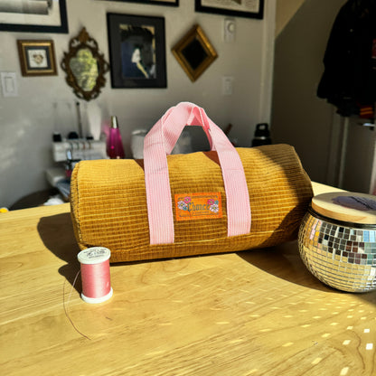 A mustard yellow corduroy cylindrical bag with pink handles sits on a wooden table in a home setting, with a spool of pink thread and a mirrored tin nearby.