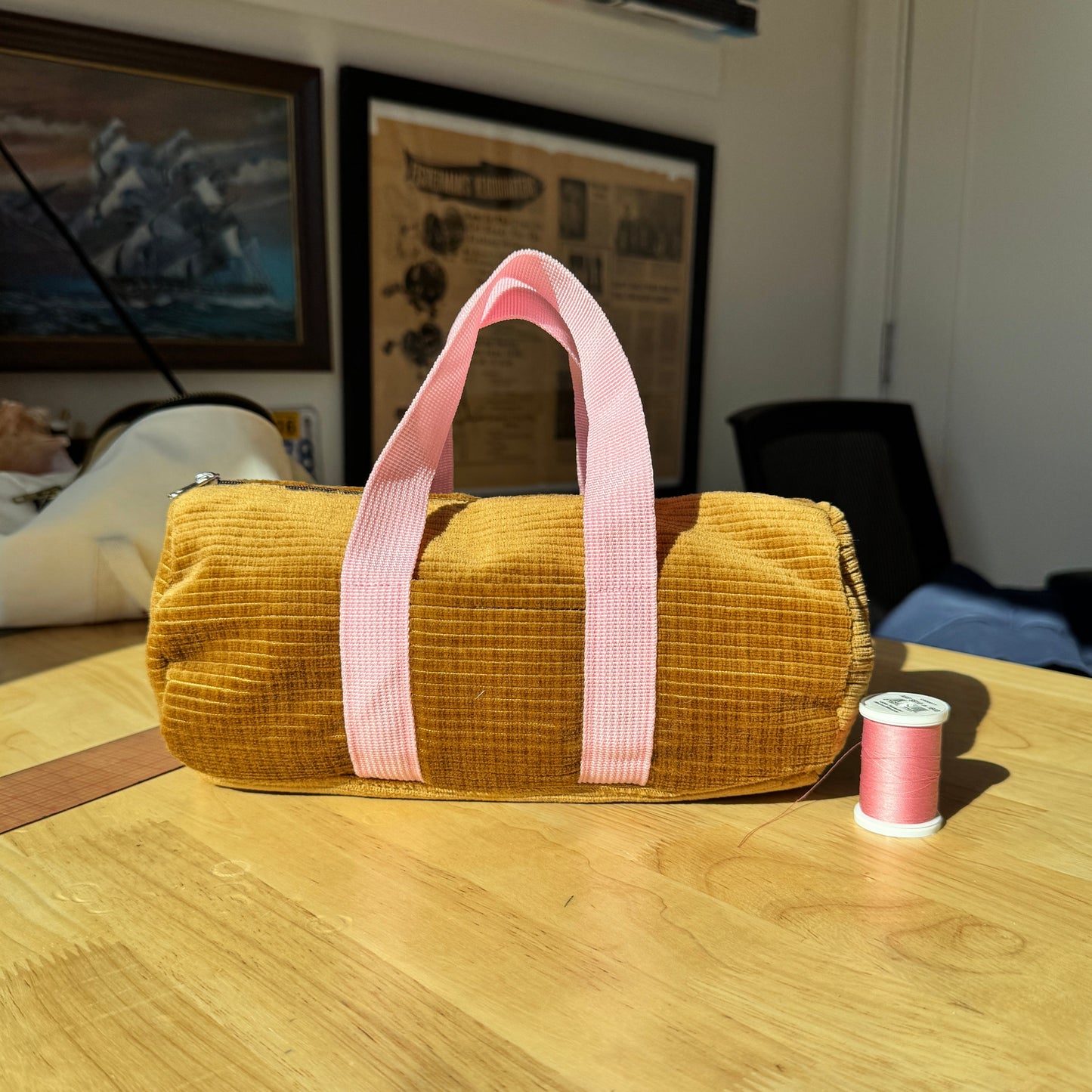 A mustard yellow corduroy cylindrical bag with pink handles sits on a wooden table in a home setting, with a spool of pink thread next to it.