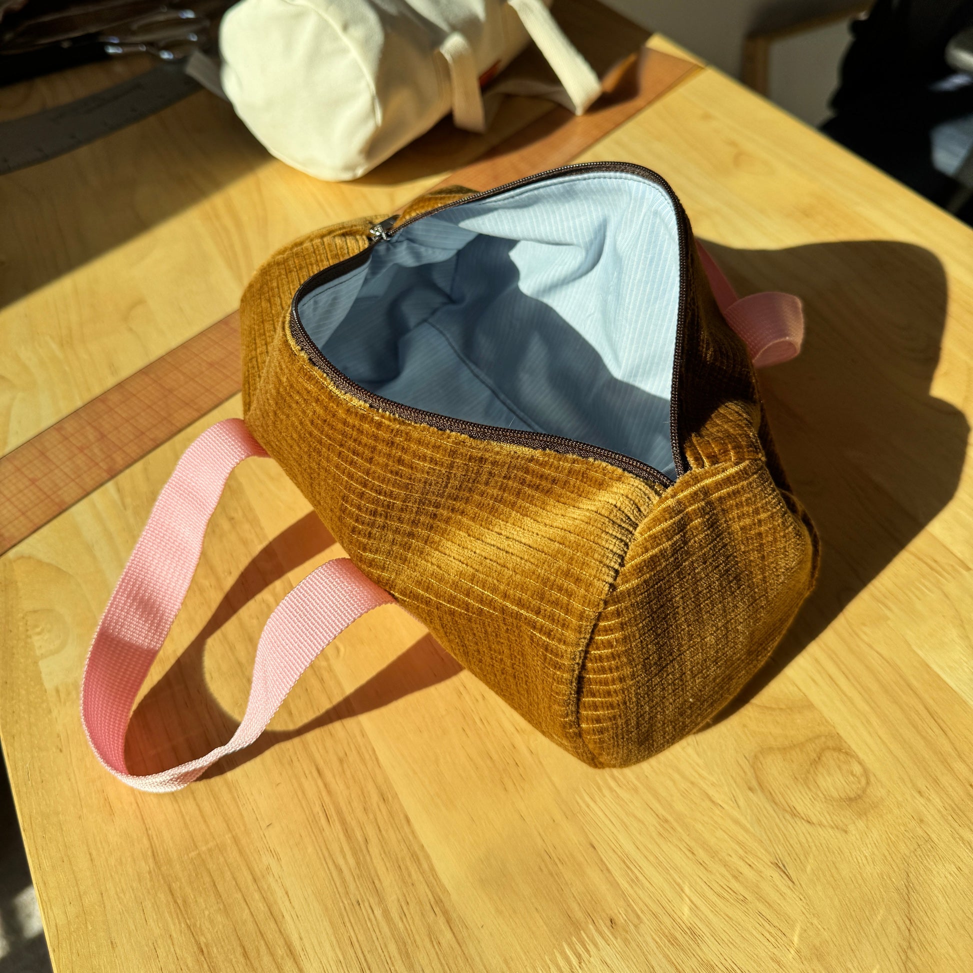 A small zippered cosmetic pouch made of textured mustard-yellow corduroy fabric, featuring a light pink fabric strap and a light blue striped interior lining, resting on a wooden table in direct sunlight.