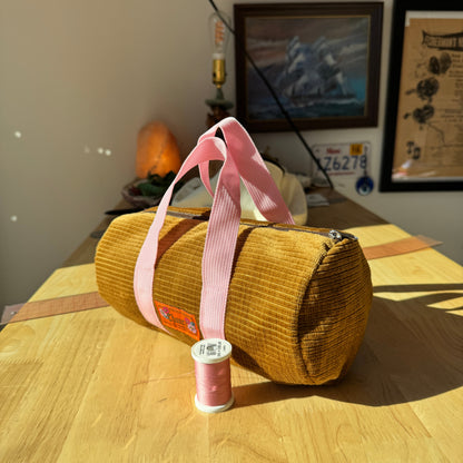 A mustard yellow corduroy cylindrical bag with pink handles sits on a wooden table in a home setting, with a spool of pink thread placed in front of it.